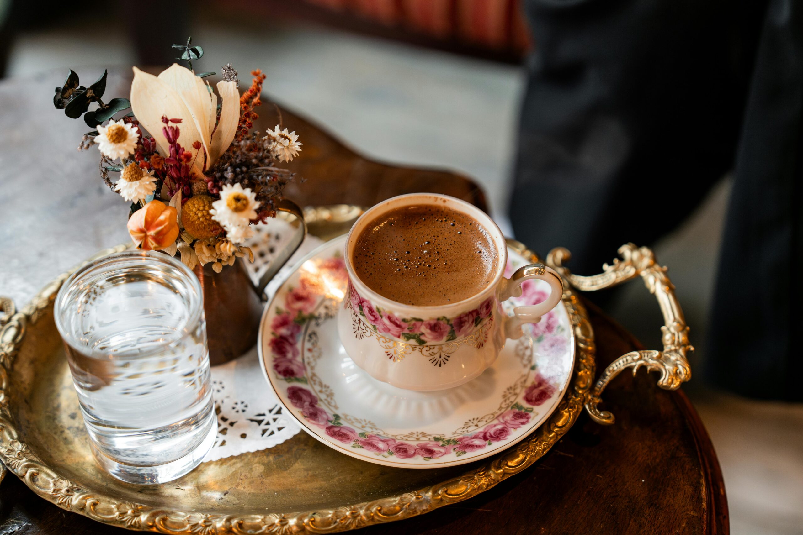 Vintage coffee setup with ornate cup, glass, and dried floral arrangement on a decorative tray.